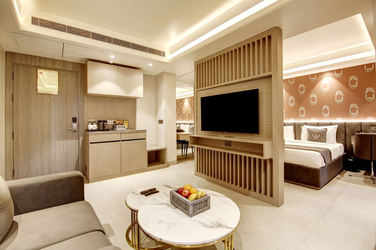 Une chambre d’hôtel moderne équipée d’une télévision à écran plat, d’un coin repas, d’un canapé, d’une petite table avec des fruits et d’un lit avec une tête de lit.