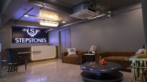 A modern reception area with a 'STEPSTONES' sign, a chandelier, a reception desk, a brown sofa, and a round coffee table with a decorative item. - Stepstones Hotels and Inn (Gurugram)