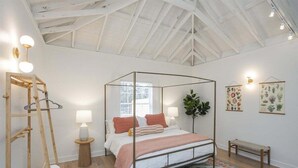 Een kamer met een hemelbed, een houten ladder, een bankje en een kamerbreed tapijt, met witte wanden en een hoge plafond.