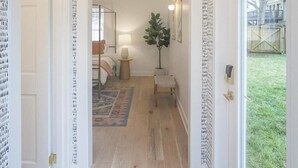 Een gang met behang, een houten vloer, een bed met vloerkleed, een plant en een raam met uitzicht op een tuin.