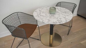 Een ronde tafel met een marmeren blad en een enkel groen glas. Twee stoelen met gaasrugleuningen en bruine kussens staan eromheen.
