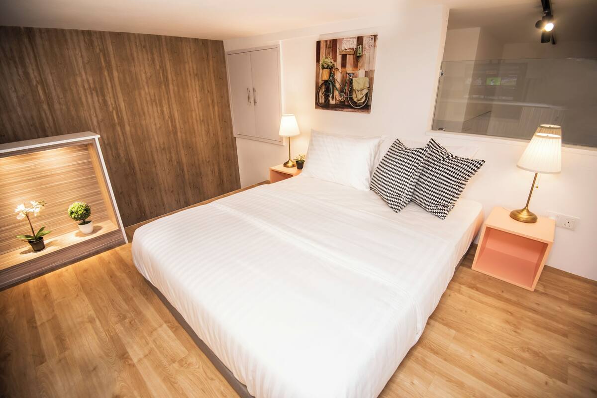 A modern bedroom with a large bed, wooden flooring, a nightstand with a lamp, a wall-mounted bike art piece, and a window with a view.