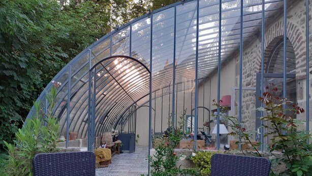 A greenhouse with a curved glass roof, wicker furniture, and potted plants.