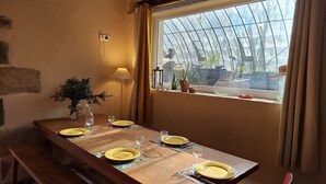 A dining area with a long wooden table set for four, a potted plant, and a view through a greenhouse window.