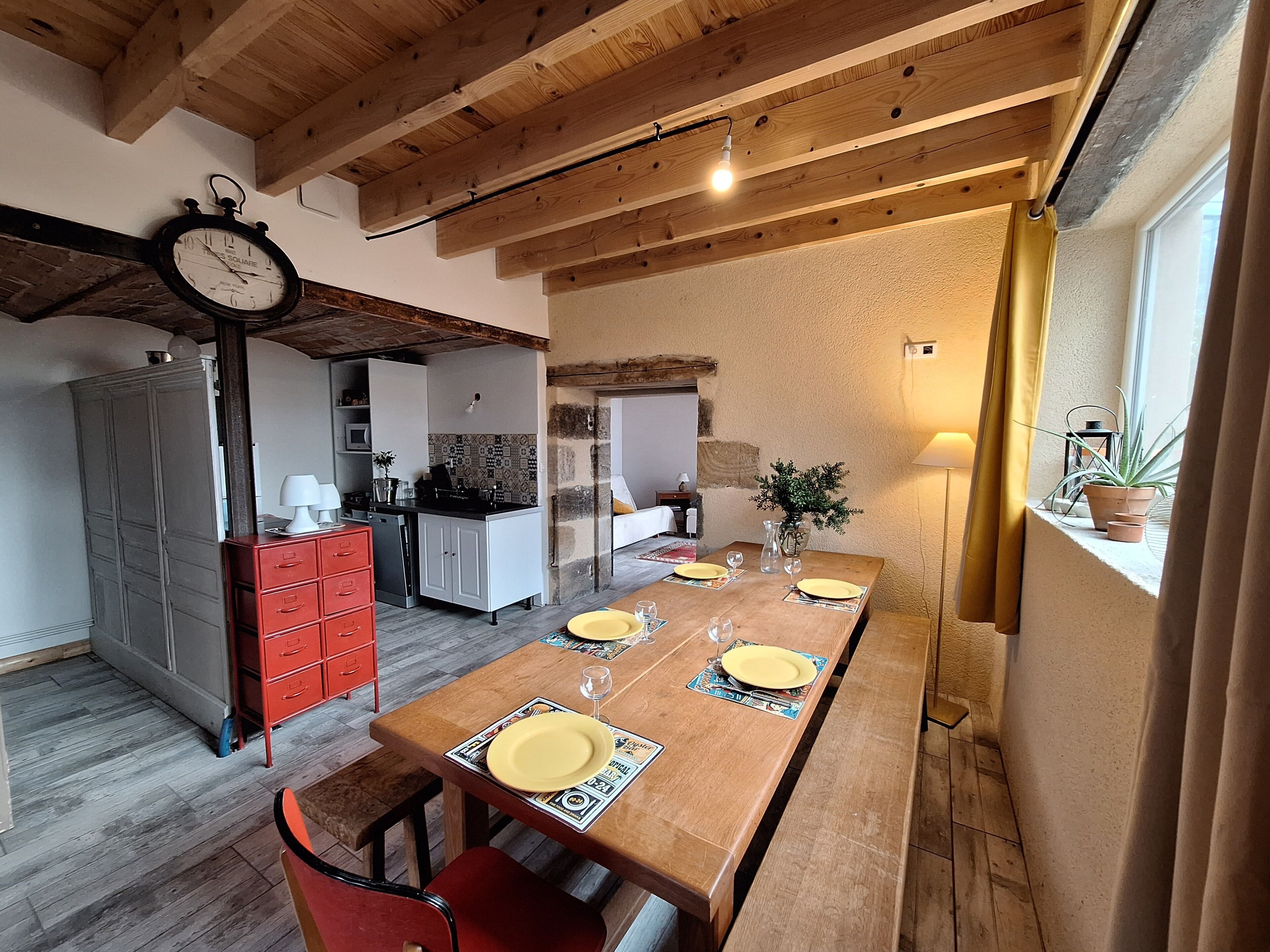 Un coin repas avec une table en bois dressée pour quatre personnes, une chaise rouge et une cuisine en arrière-plan.