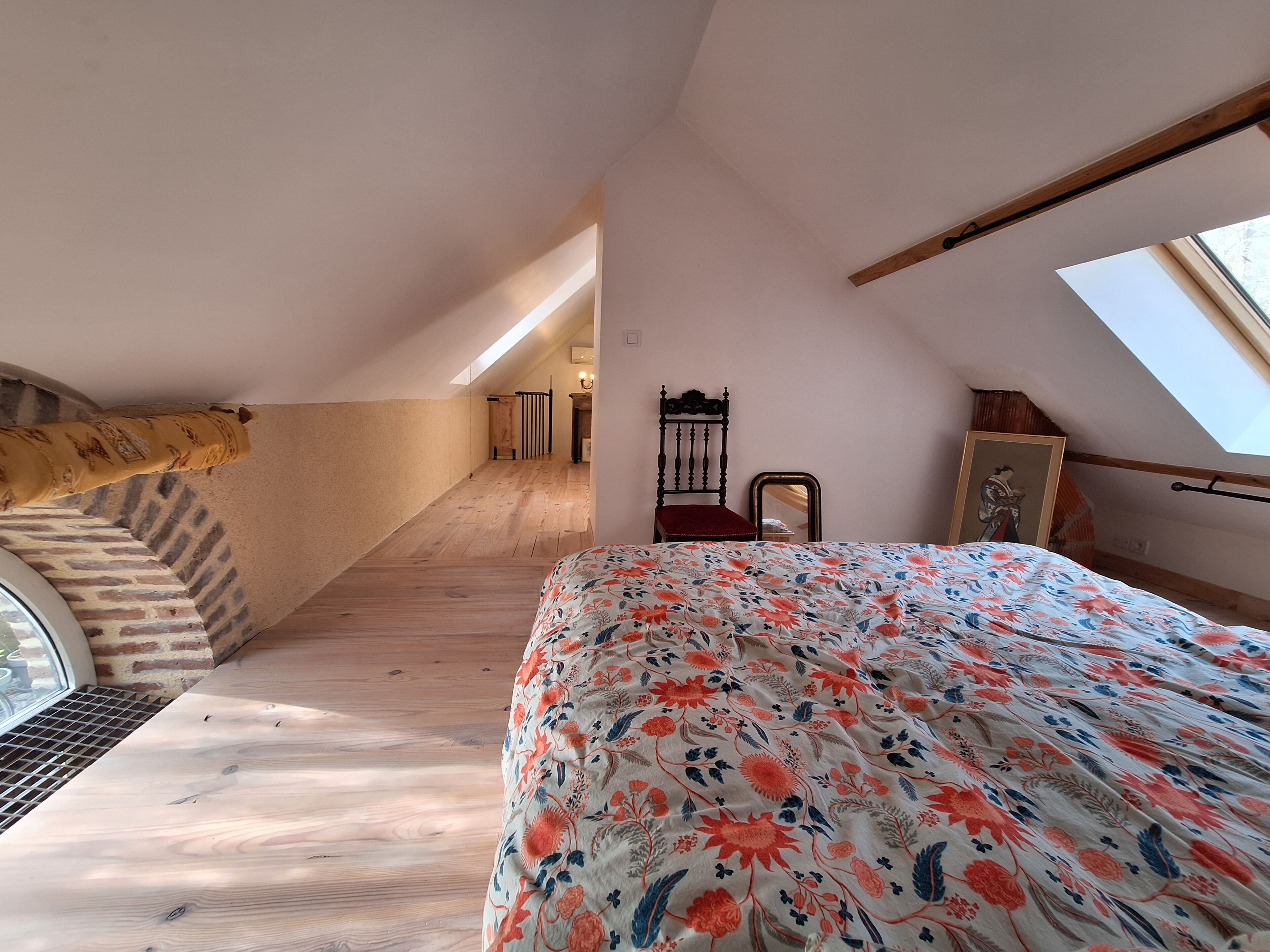 Un dormitorio con techo inclinado, una escalera de madera, una cama con colcha floral, una silla y un cuadro en la pared.