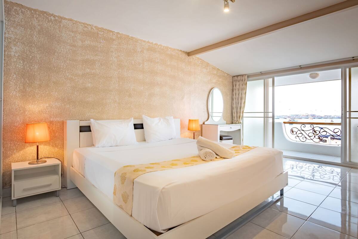 A modern hotel room with a large bed, two bedside lamps, a nightstand, a dresser, and a large window with a balcony.