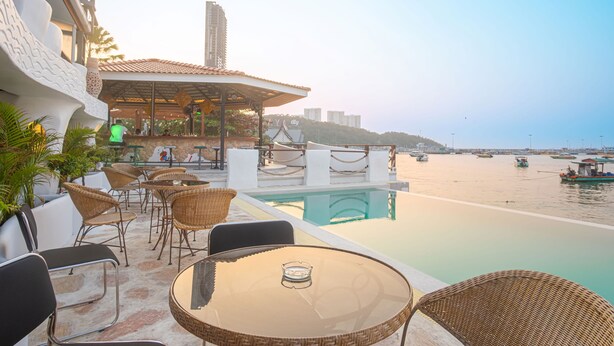 An outdoor seating area with wicker chairs and tables, a pool, and a view of boats on the water. - Windy Inn (Pattaya)
