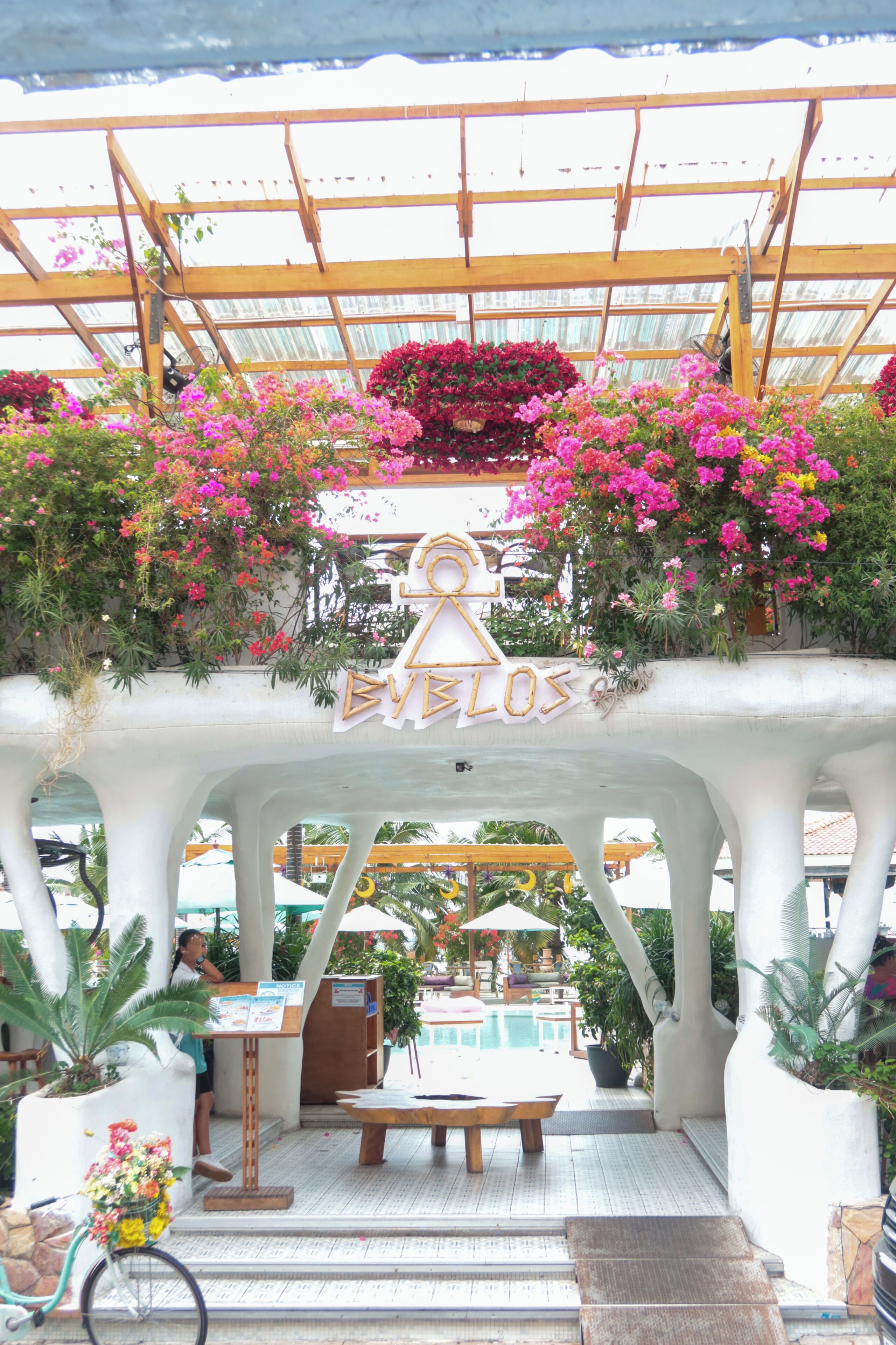 A covered outdoor seating area with a pergola, a pool in the background, and a sign that reads 'EVELOS'.