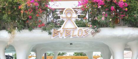 A covered outdoor seating area with a pergola, a pool in the background, and a sign that reads 'EVELOS'.