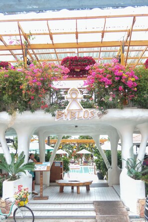 A covered outdoor seating area with a pergola, a pool in the background, and a sign that reads 'EVELOS'.
