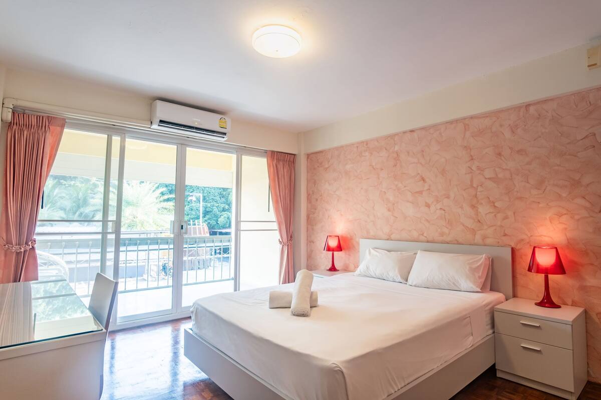A hotel room with a large bed, two bedside tables with lamps, a window with curtains, and an air conditioning unit.