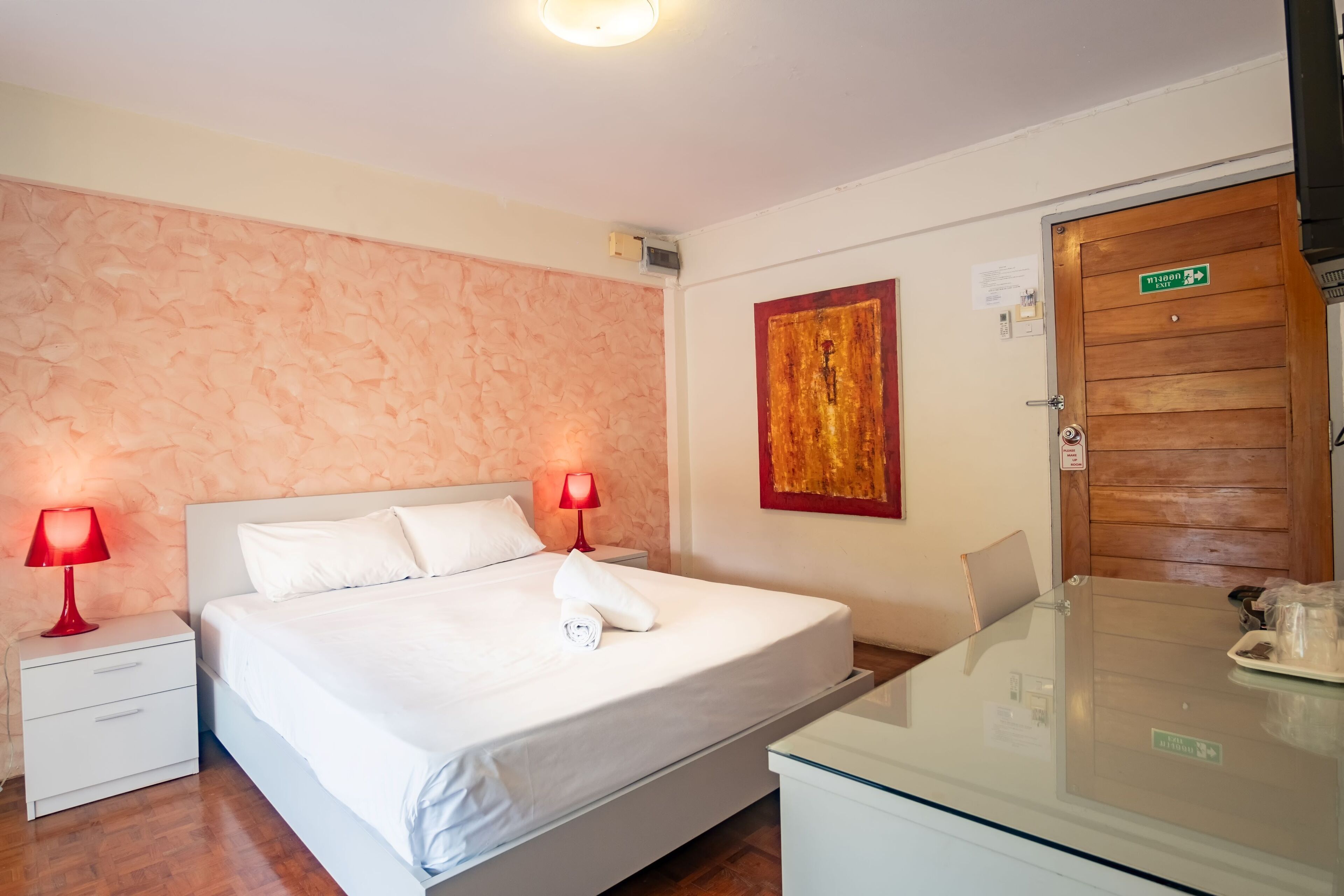 Habitación de hotel con cama, dos mesitas de noche con lámparas, una puerta de madera y una mesa de cristal con un cartel.