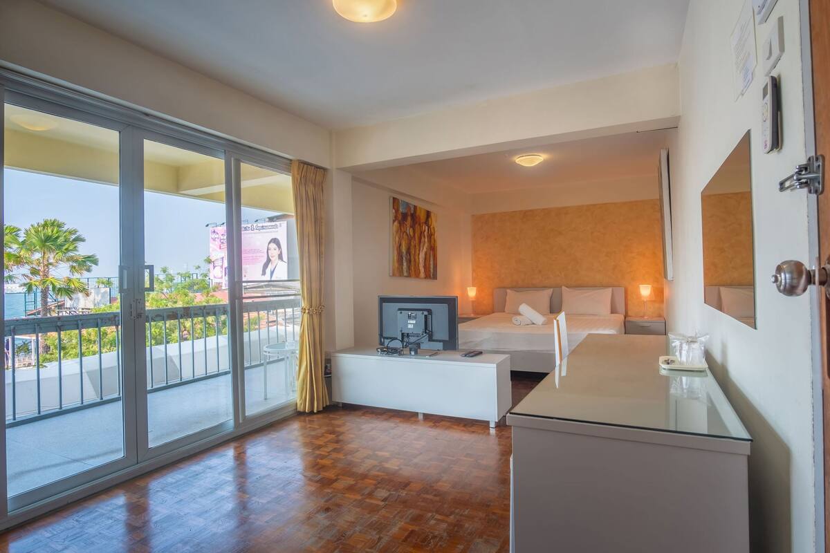 A hotel room with a large sliding glass door to the balcony, a bed with white linens, a small desk with a television, and a bedside table with a lamp.