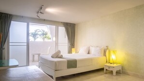 A hotel room with a bed, a nightstand with a lamp, a small table, and a view of the outdoors through a large window. - Windy Inn (Pattaya)