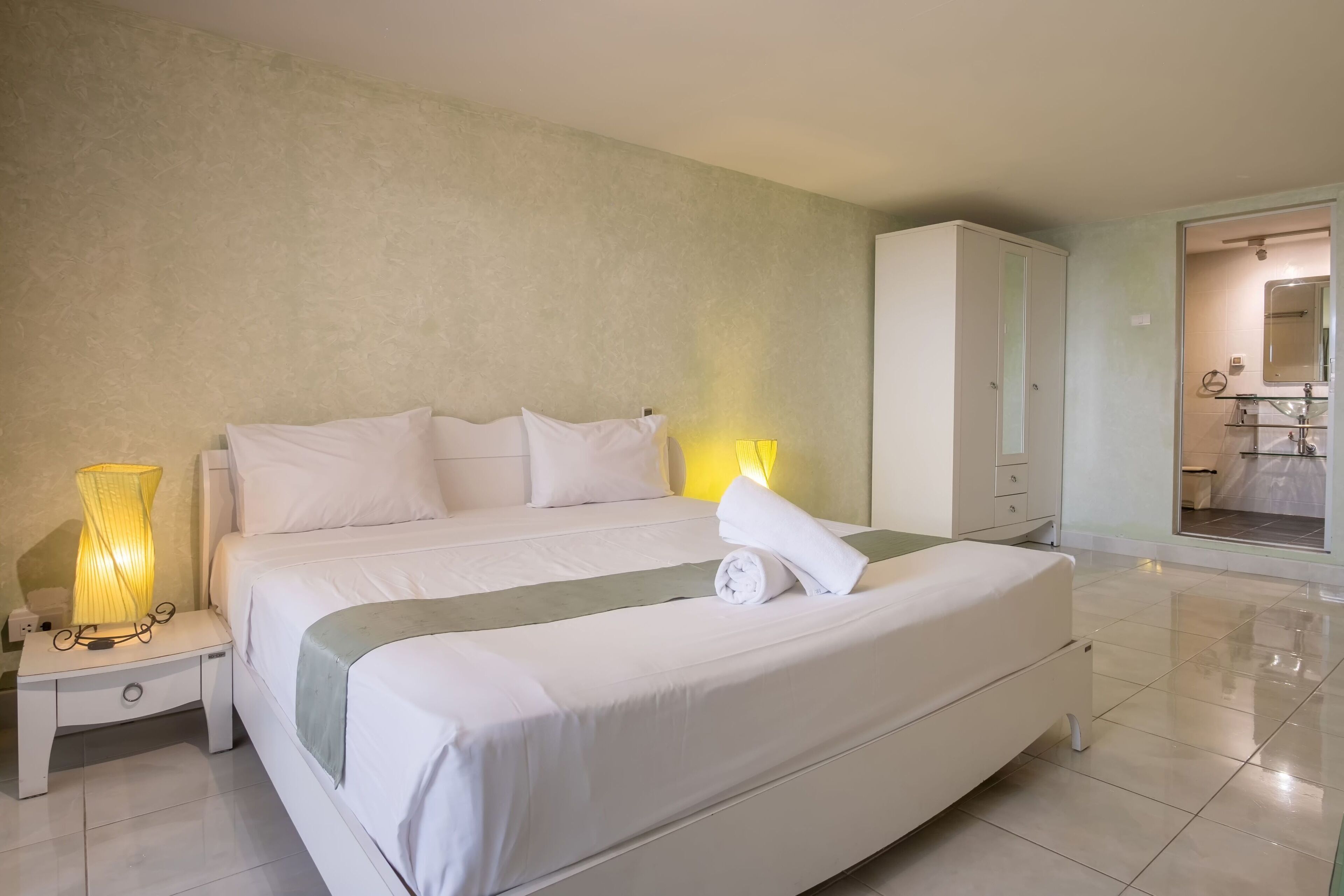 A neatly made bed with white linens and a rolled towel, a bedside lamp, and a wardrobe in a hotel room.