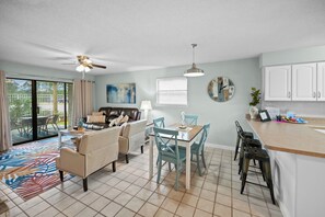 A spacious living area with a ceiling fan, a dining table with chairs, a sofa, and a kitchen area with cabinets and a countertop. - Ground Floor Unit, Seaside Beach and Racquet Club - Signature Properties (Orange Beach)