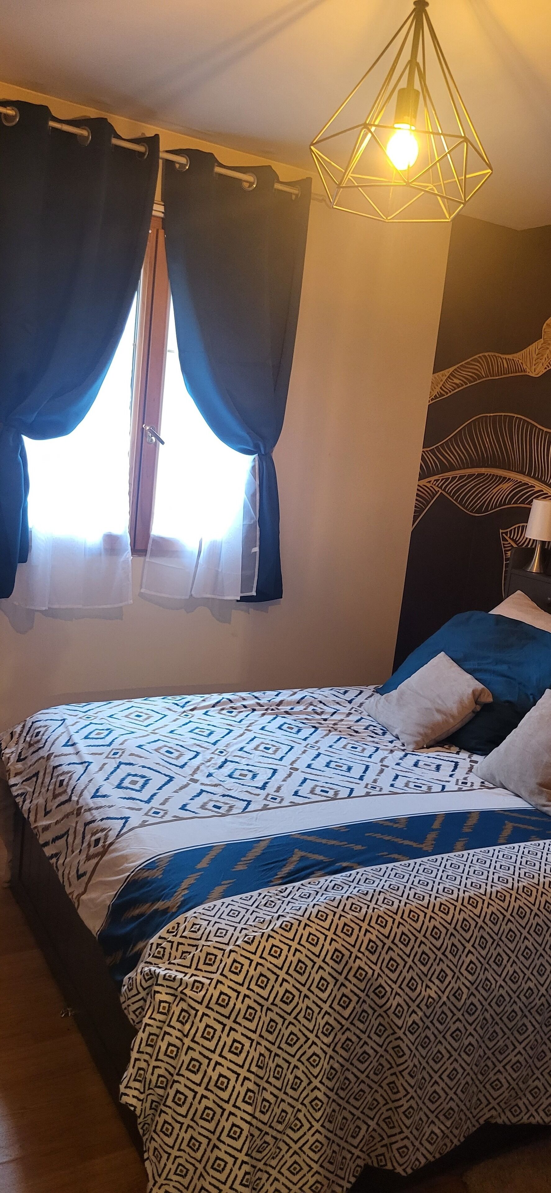 A bedroom with a bed, curtains, a hanging light fixture, and a patterned wall.