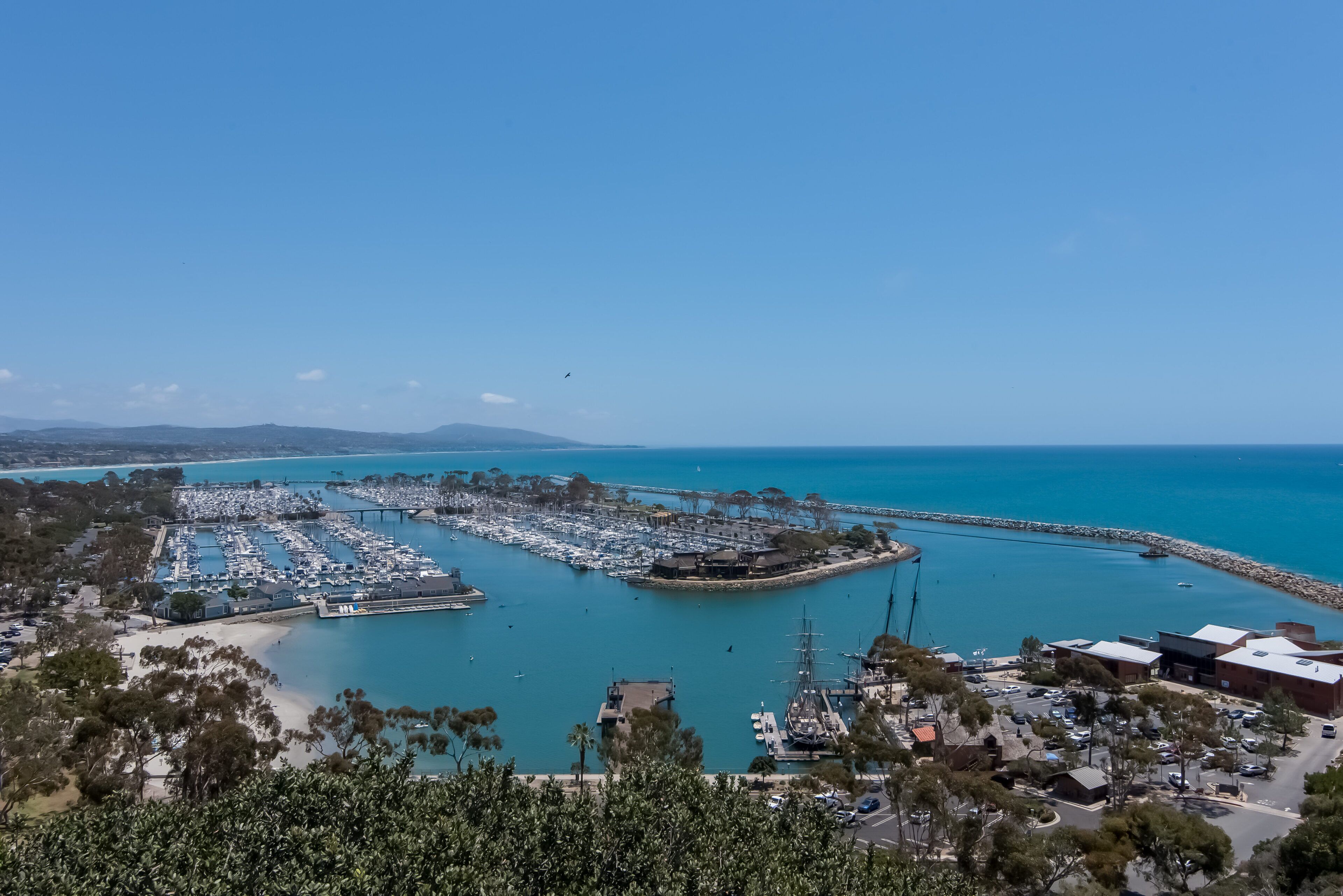Vue aérienne d’une marina avec des bateaux amarrés, une jetée et des bâtiments offrant une vue dégagée sur le paysage environnant.