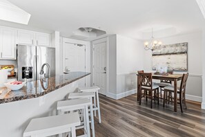 A modern kitchen with white cabinets, a stainless steel refrigerator, and a granite countertop. There is a dining area with a wooden table and chairs.