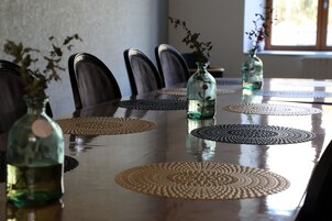 Una mesa de comedor con superficie reflectante, rodeada de sillas y decorada con individuales tejidos y jarrones de vidrio con flores secas.