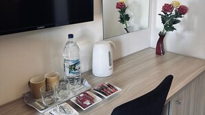 Une chambre d’hôtel avec un bureau en bois, un téléviseur à écran plat, un miroir, un vase avec des fleurs, une bouilloire, une bouteille d’eau, une télécommande et quelques magazines.