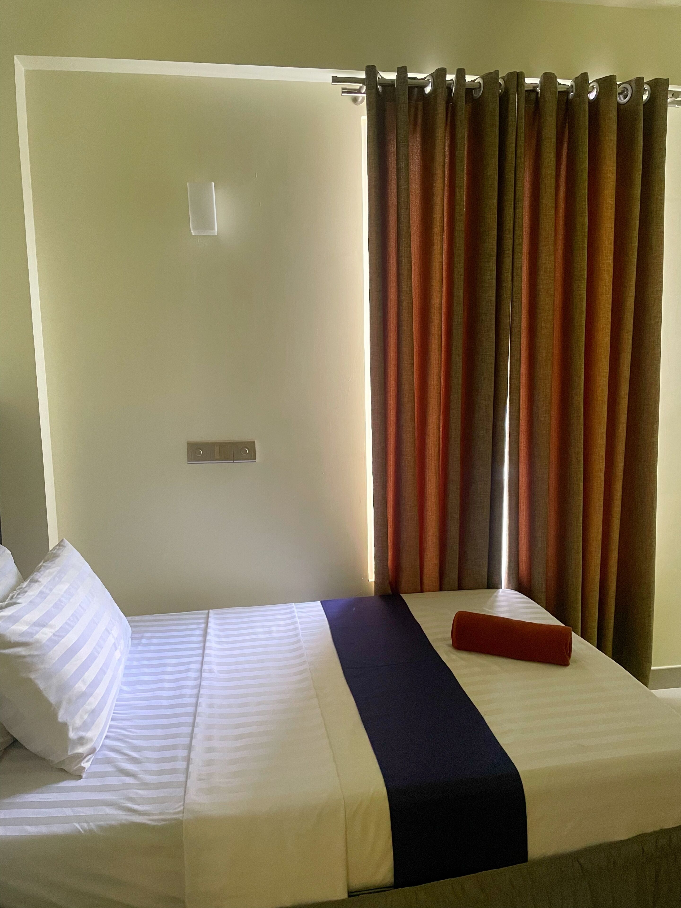 Habitación de hotel con cama, almohada roja, almohada a rayas, corredor azul oscuro para la cama y una cortina.