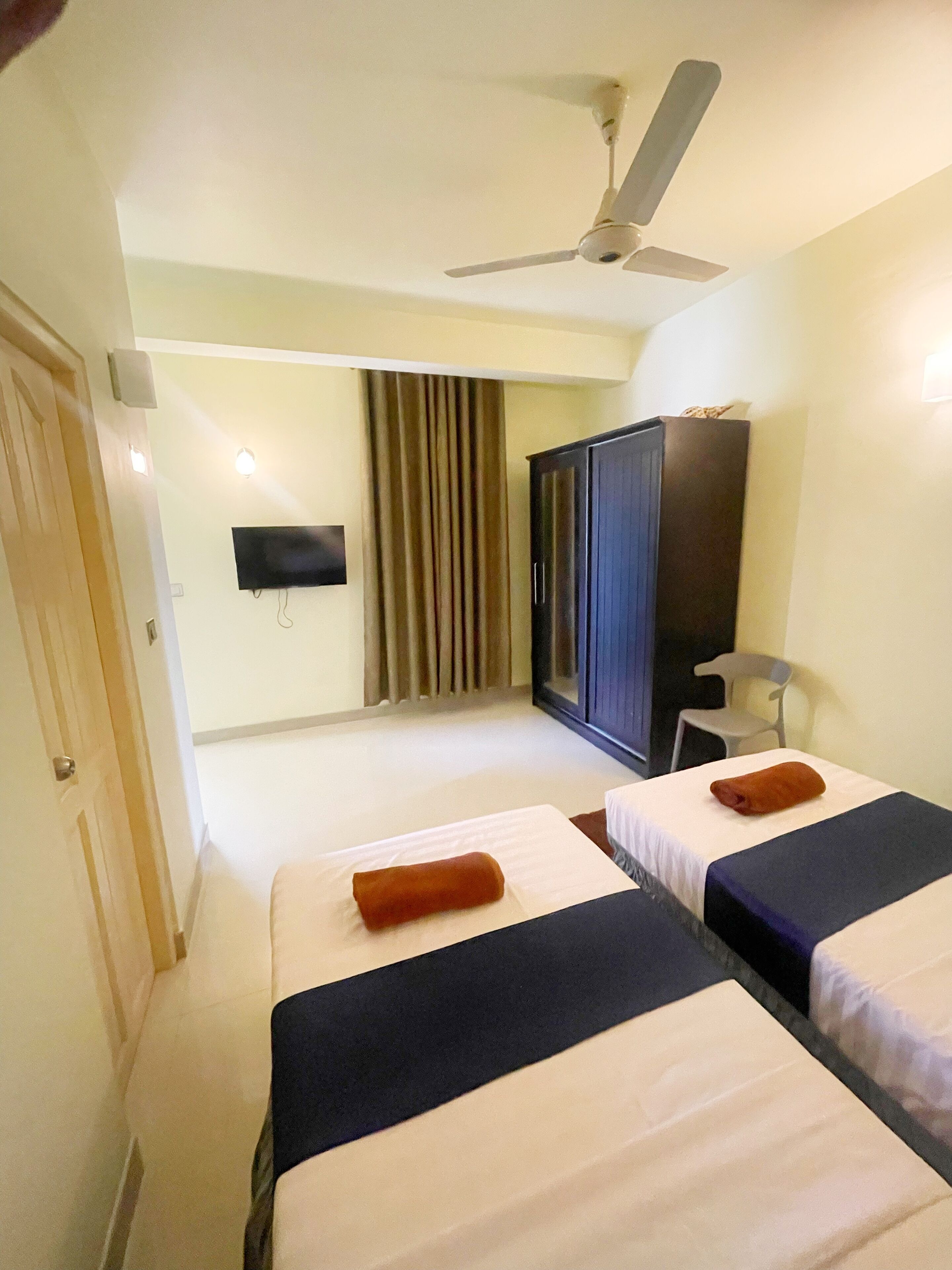 A hotel room with two beds, a ceiling fan, a TV mounted on the wall, and a wardrobe.