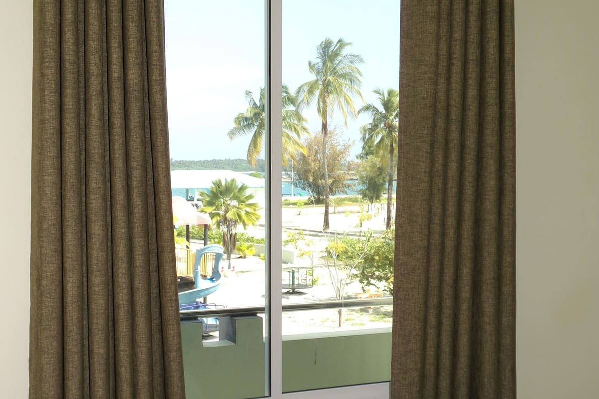 A hotel room with a bed, curtains, and a window with a view of palm trees and a pool.