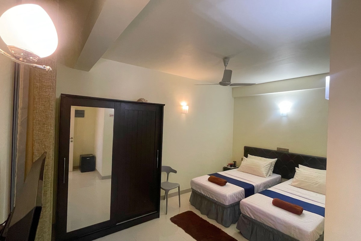 A hotel room with two beds, a wardrobe, a mirror, a ceiling fan, and a small desk.