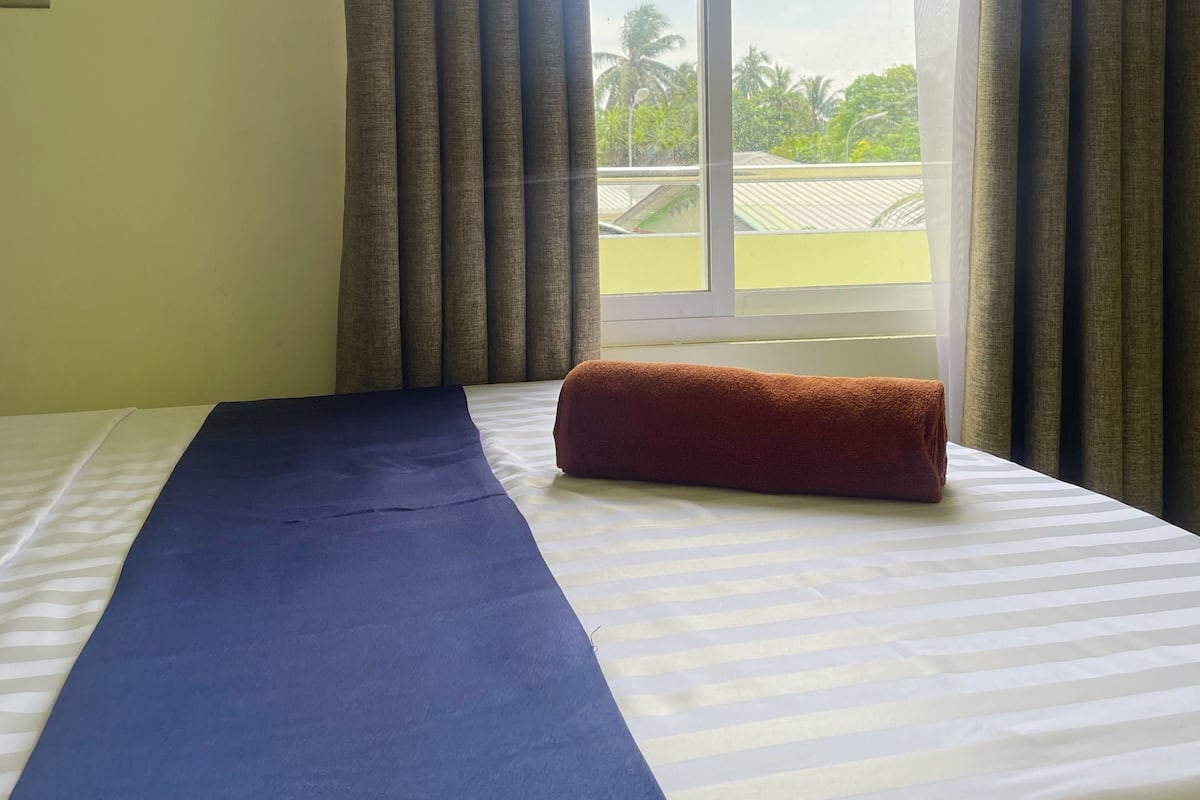 A hotel room with a bed, a window with curtains, and a view of greenery outside.