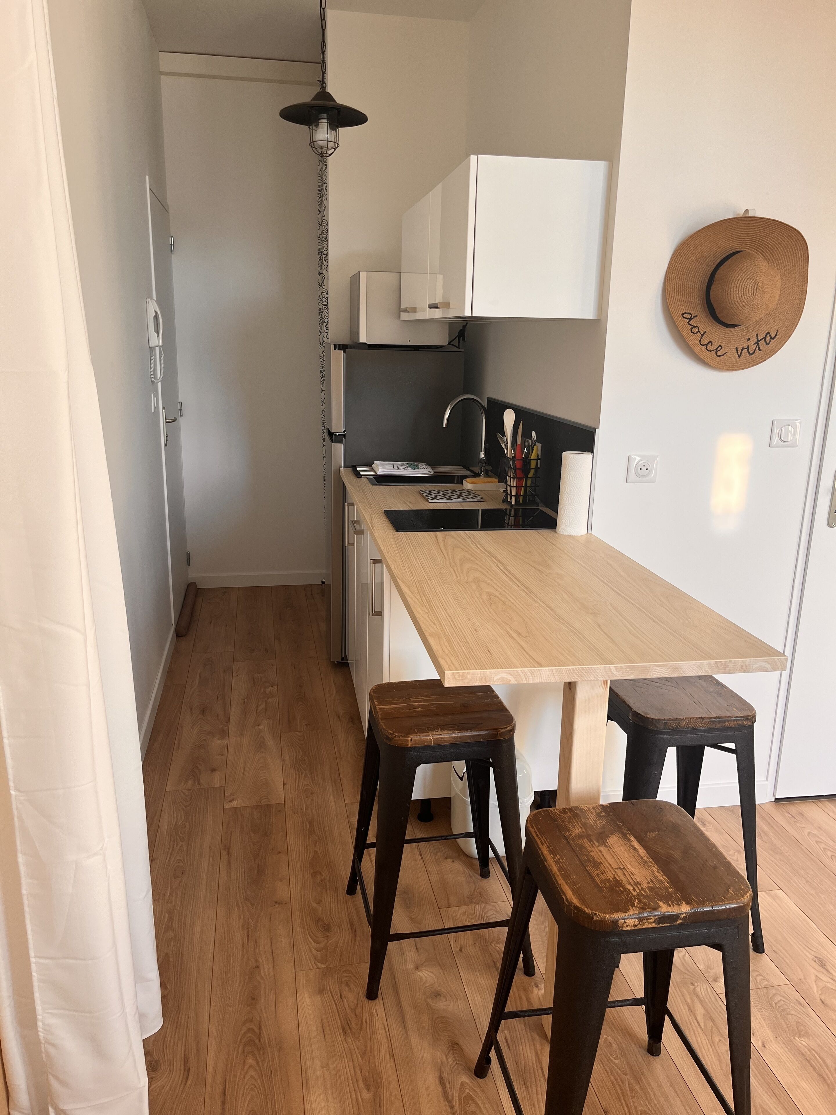Una cocina pequeña con una mesa de madera, dos taburetes, una lámpara colgante y un sombrero en la pared.
