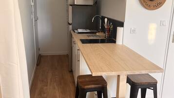 Una cocina pequeña con una mesa de madera, dos taburetes, una lámpara colgante y un sombrero en la pared.