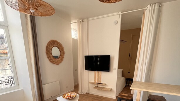 A modern living room with a sofa, a dining table with chairs, a TV mounted on the wall, and a large woven light fixture. - Appartement Élégant (Angoulême)