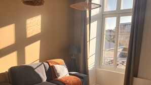 A modern living room with a sofa, a coffee table, and a dining table. There is a window with curtains and a view of the outside. - Appartement Élégant (Angoulême)