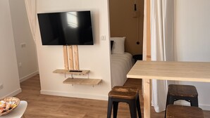 A room with a TV mounted on the wall, a wooden table, two stools, a basket on a small table, and a bed with white bedding. - Appartement Élégant (Angoulême)
