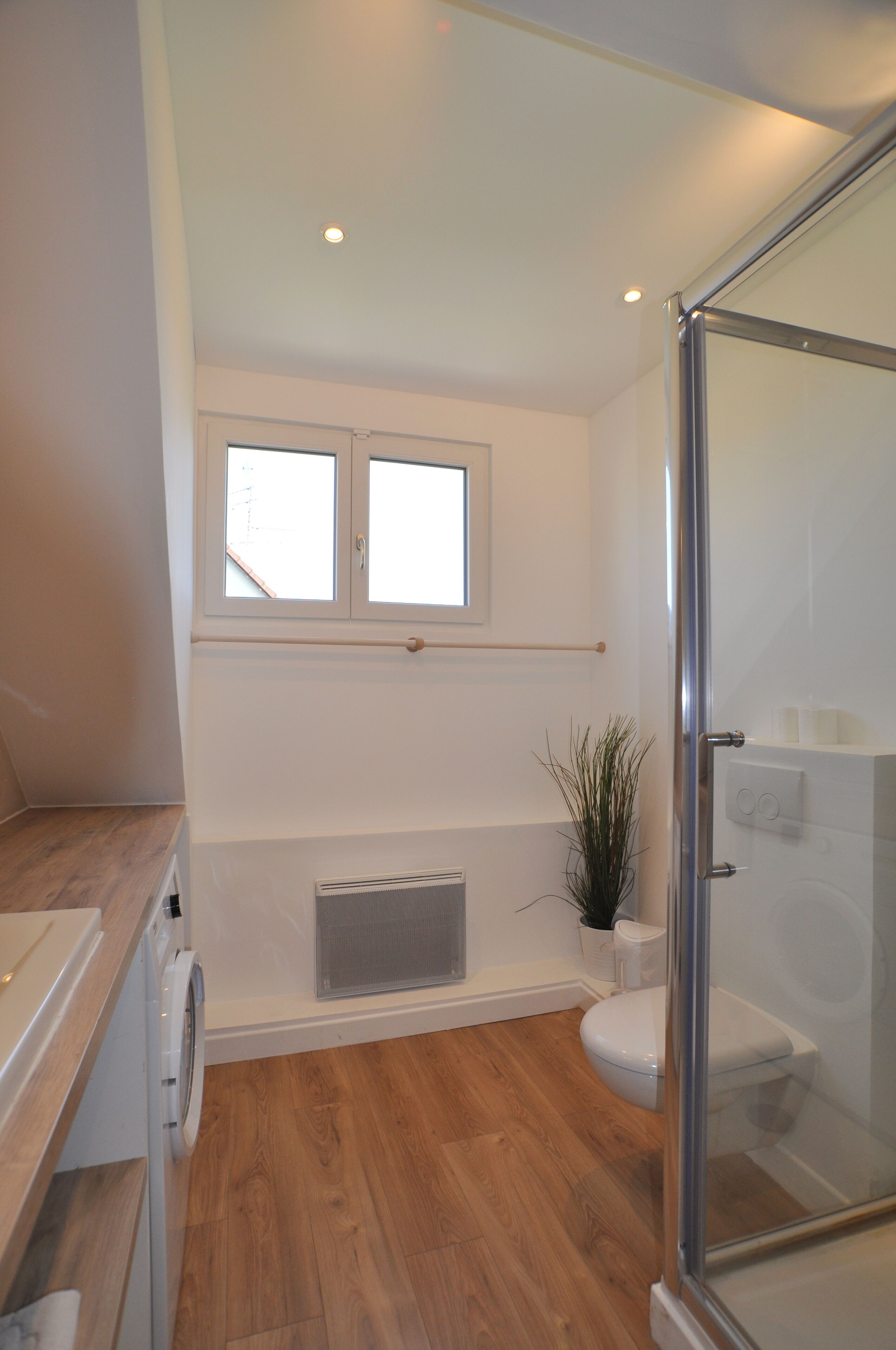 Een moderne badkamer met een glazen douchecabine, een raam boven de wastafel en een houten vloer.