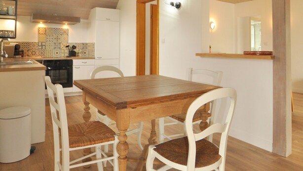 A dining area with a wooden table and white chairs, a kitchen in the background, and a sloped ceiling with exposed beams.