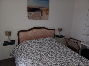 A bedroom with a bed, two bedside tables with lamps, a chair, a shelf, and a painting of a beach scene. - Maison Avec vue mer (Lanildut)