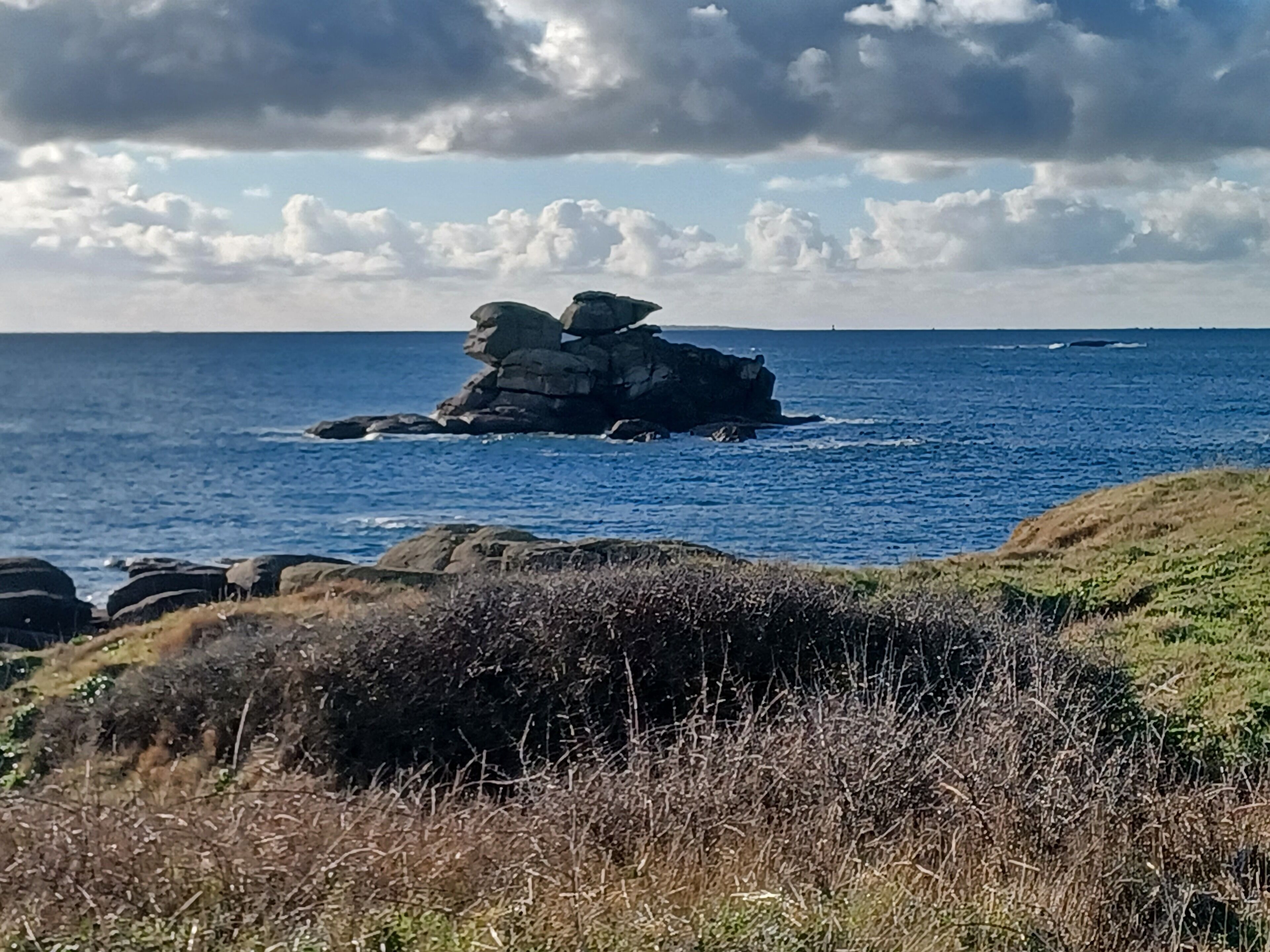 Un paysage côtier avec une formation rocheuse imposante qui s’élève au-dessus de la mer, entourée d’une étendue herbeuse et sous un ciel dégagé.