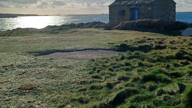Een stenen gebouw met een blauwe deur en schoorsteen, gelegen op een grasveld met uitzicht op zee.