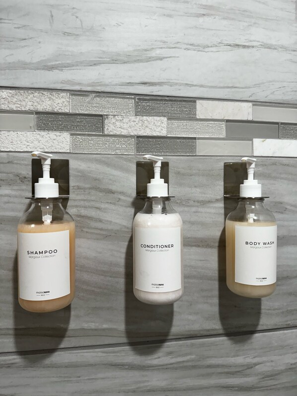 Three bottles of shampoo, conditioner, and body wash from the Margaux Collection, hanging on a tiled wall.