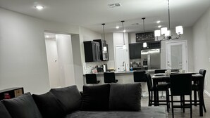 A modern living room with a sectional sofa, a dining area with a table and chairs, a kitchen with dark cabinets and stainless steel appliances, and a ceiling fan.