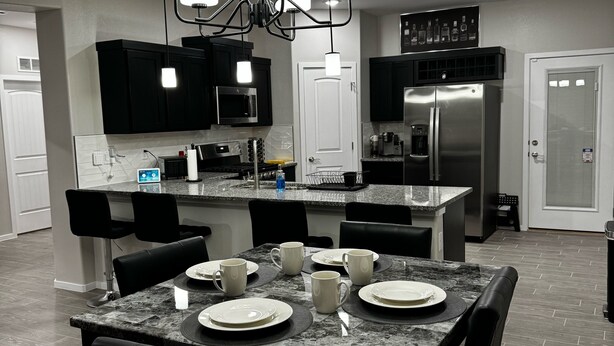 A modern kitchen with a central island, black cabinets, stainless steel appliances, and a chandelier.