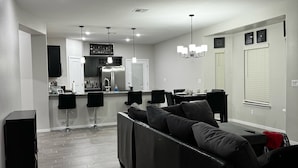 A modern living room with a black sectional sofa, a dining area with a table and chairs, and a kitchen in the background.
