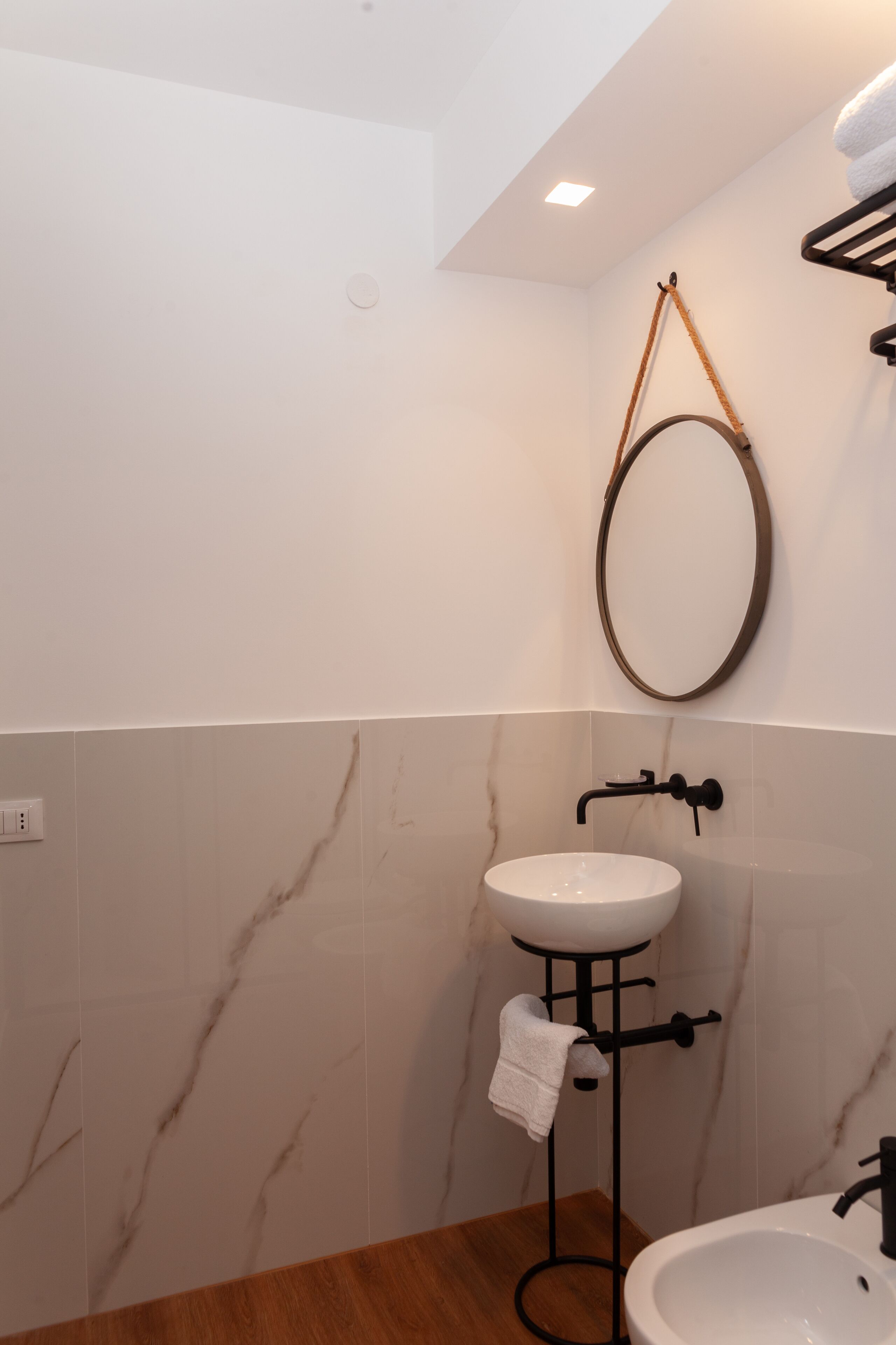A bathroom with a marble wall, a pedestal sink, a round mirror, and a towel rack.