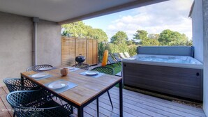 Een terras buiten met een houten tafel en stoelen, een jacuzzi en uitzicht op bomen.