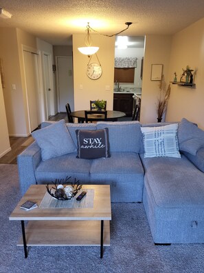 A living room with a gray sectional sofa, a wooden coffee table, and a dining area in the background. - Cozy condo, walking distance to trails, shops, restaurants, fitness center.
 (Grand Junction)