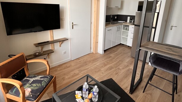 A modern living room with a flat-screen TV, wooden chairs, a coffee table with snacks, a black hanging light fixture, and a kitchen area in the background. - Charmant Appartement (Angoulême)