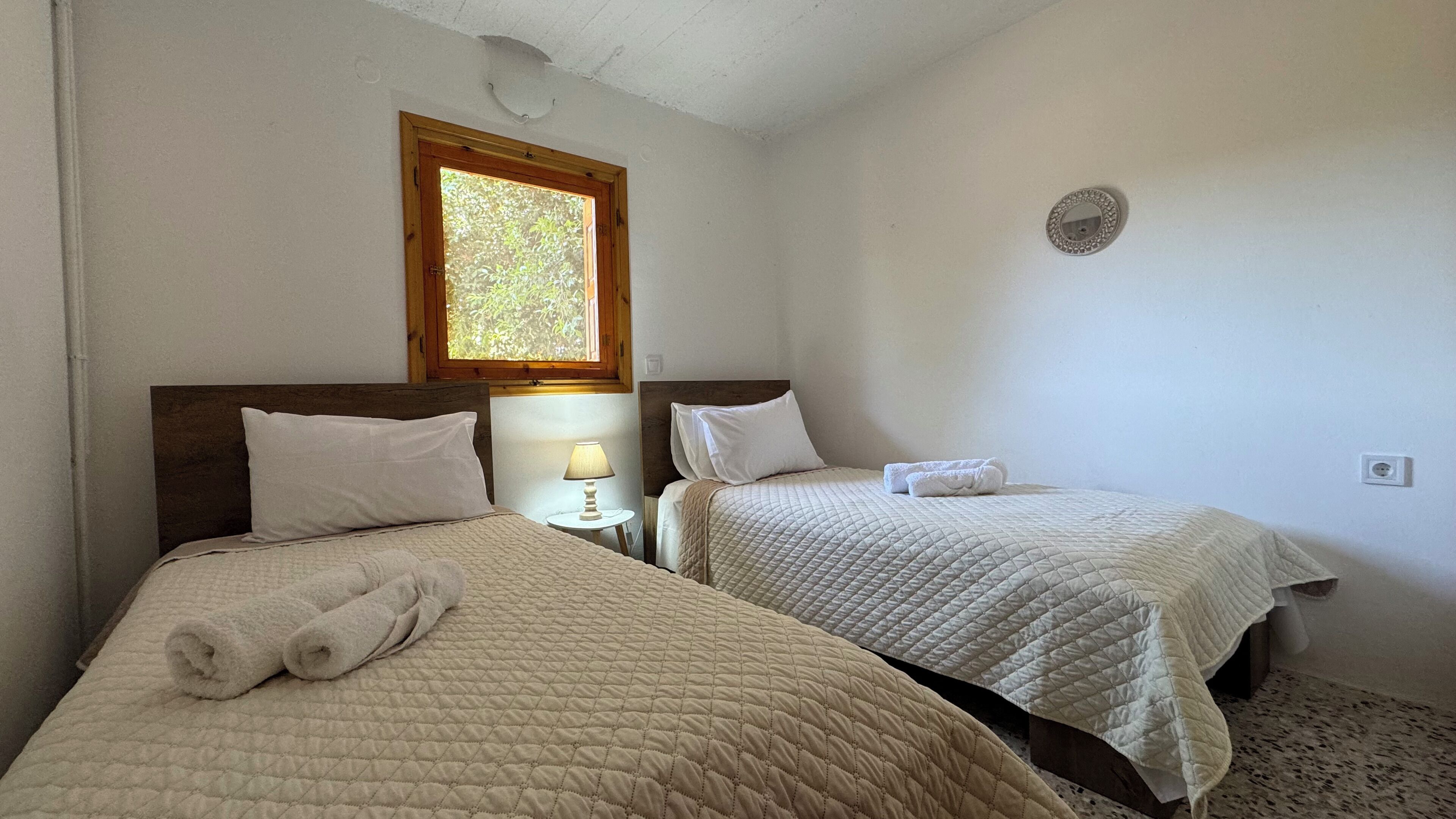 Ein Zimmer mit zwei Betten, jeweils mit gesteppter Bettdecke und Kissen, einem Nachttisch mit Lampe und einem Fenster mit Blick auf Bäume.